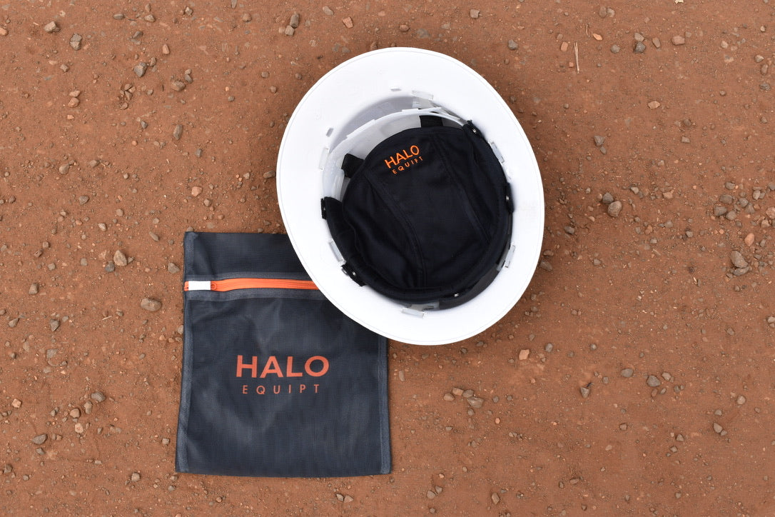 Black HALO Lid Liner fitted inside a white hard hat, beside the black HALO Wash Bag on red dirt.