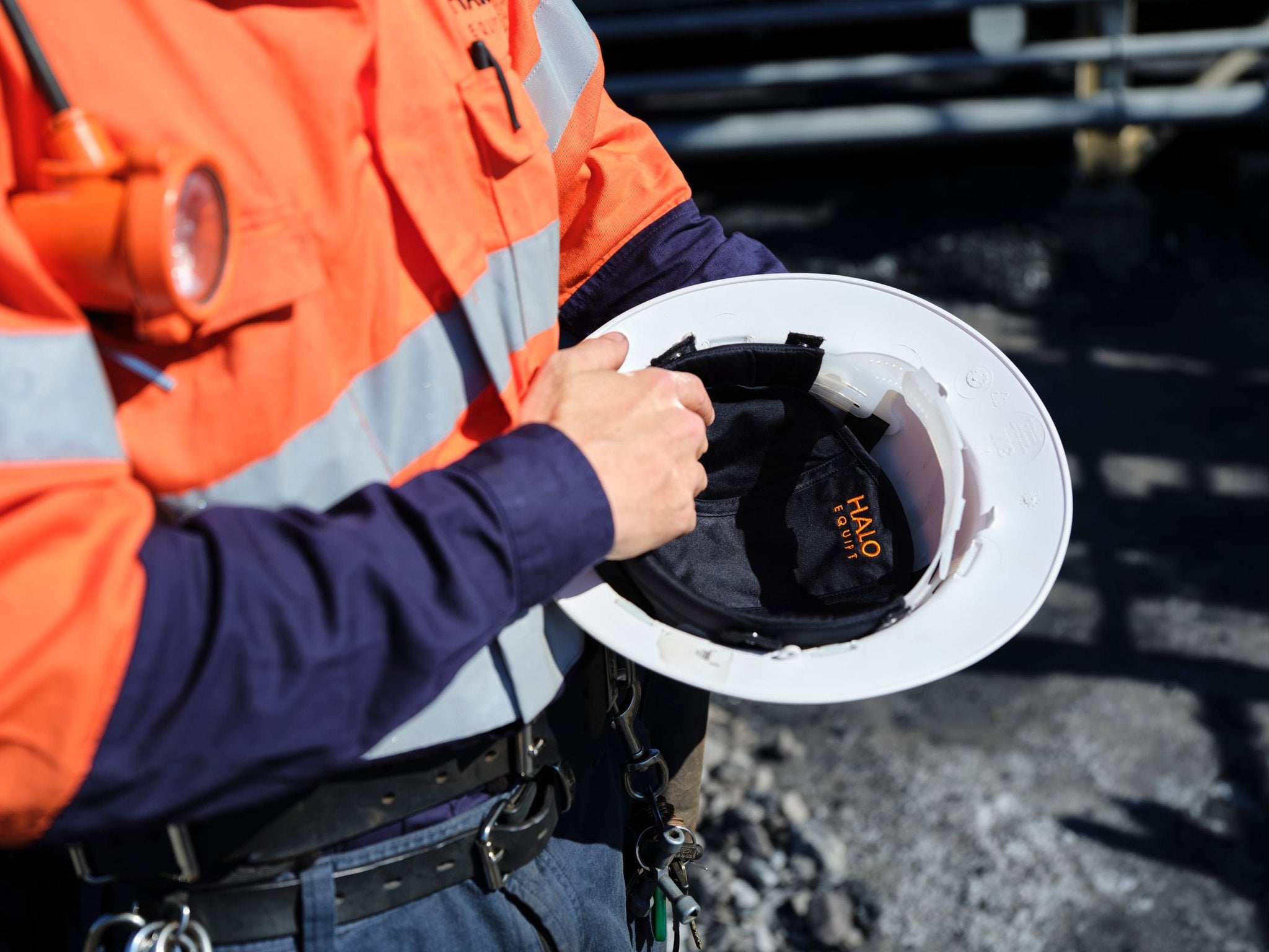 A worker in hi-vis holding a white hard hat open on a coal handling site, a black HALO Lid Liner fitted inside with the orange Halo Equipt embroidery showing