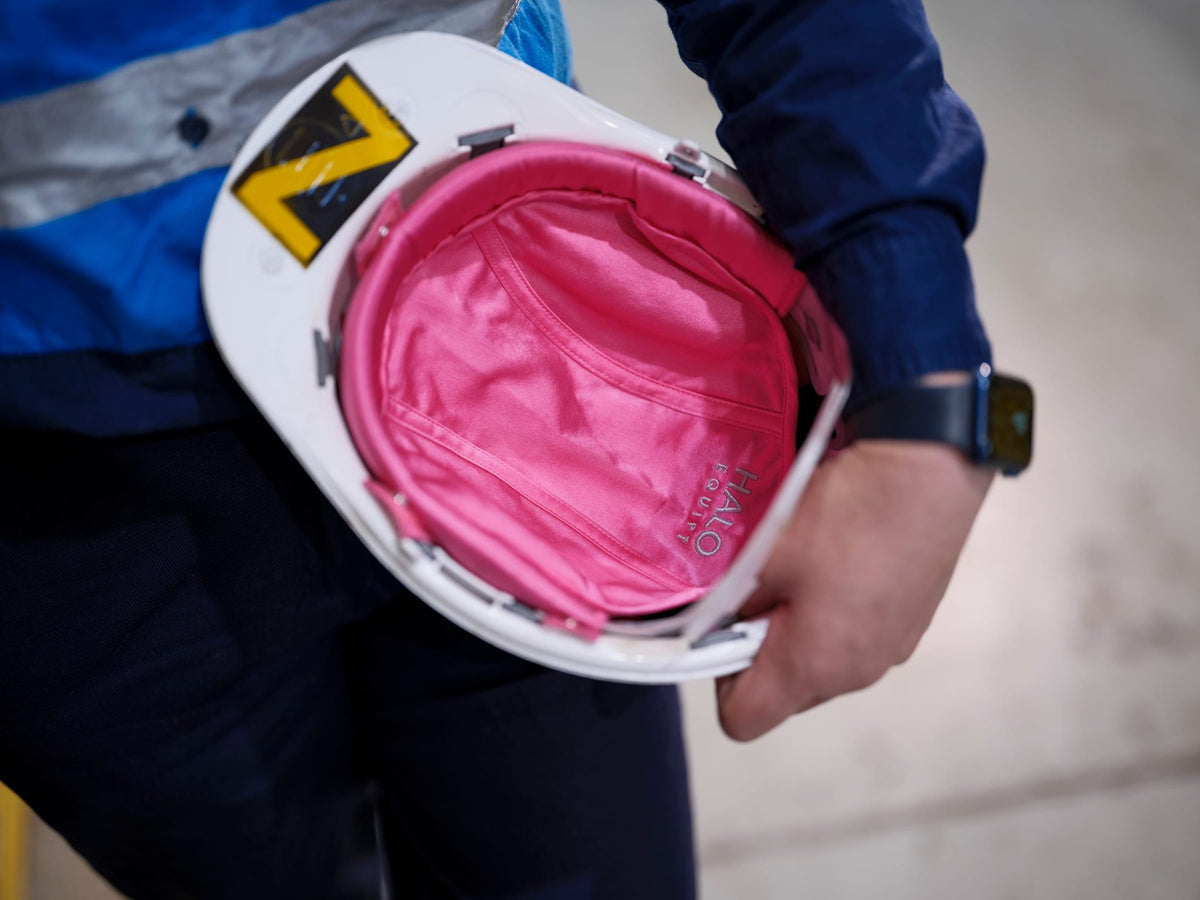A pink HALO Lid Liner fitted inside a white hard hat, held at the wearer side on a worksite