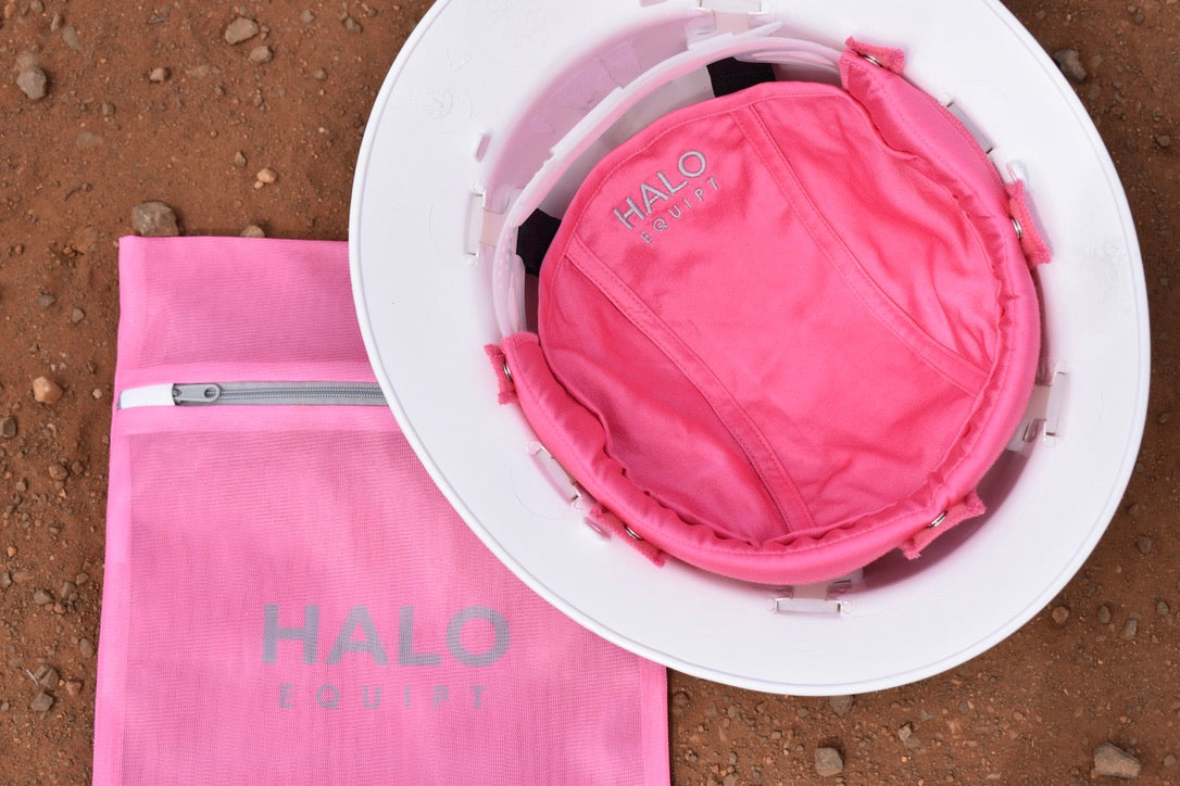 Pink HALO Lid Liner fitted inside a white hard hat, beside the pink HALO Wash Bag on red dirt.
