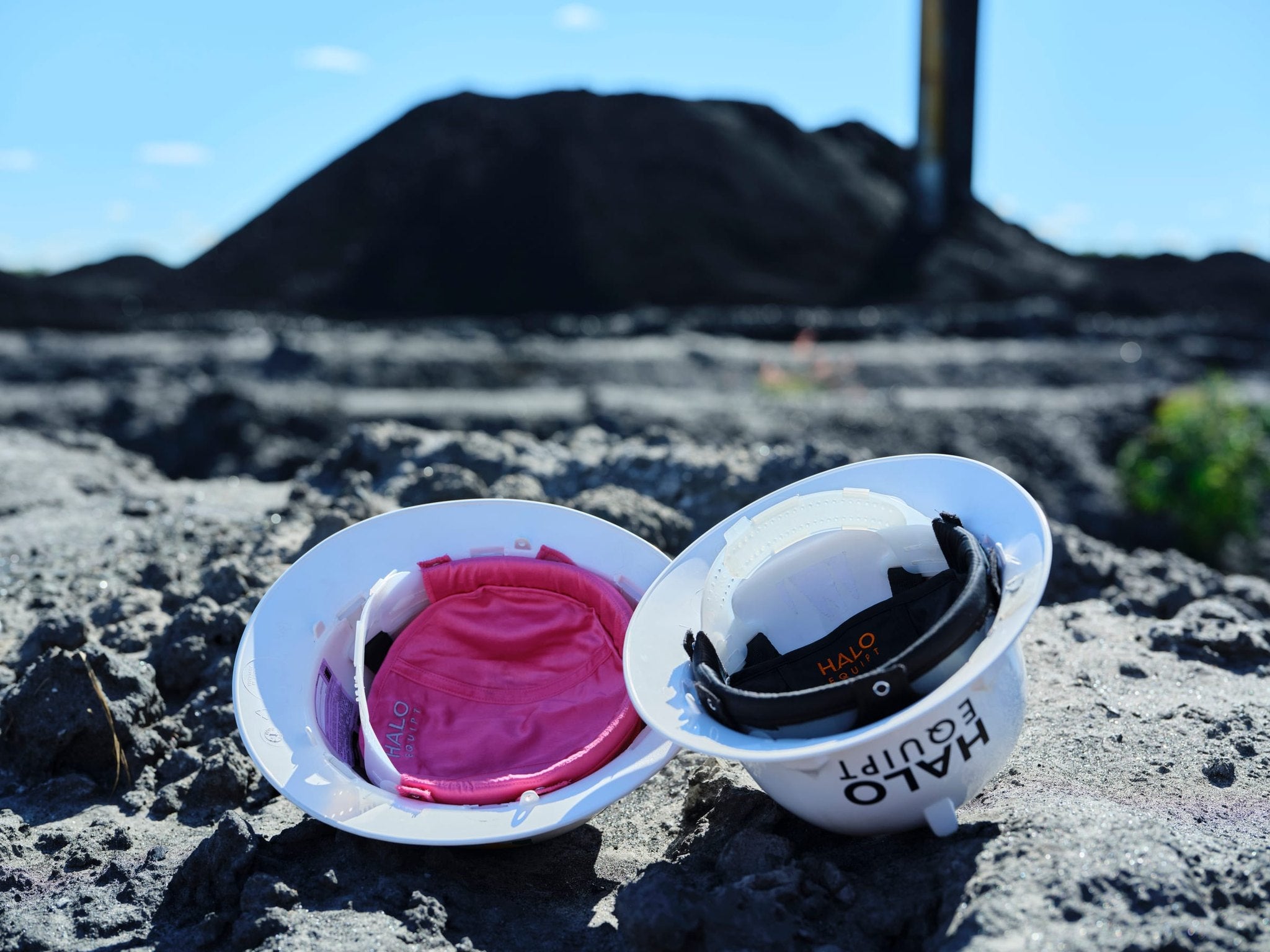 Two white hard hats resting on rock at a coal stockpile under blue sky, one fitted with a pink HALO Lid Liner and one with a black liner
