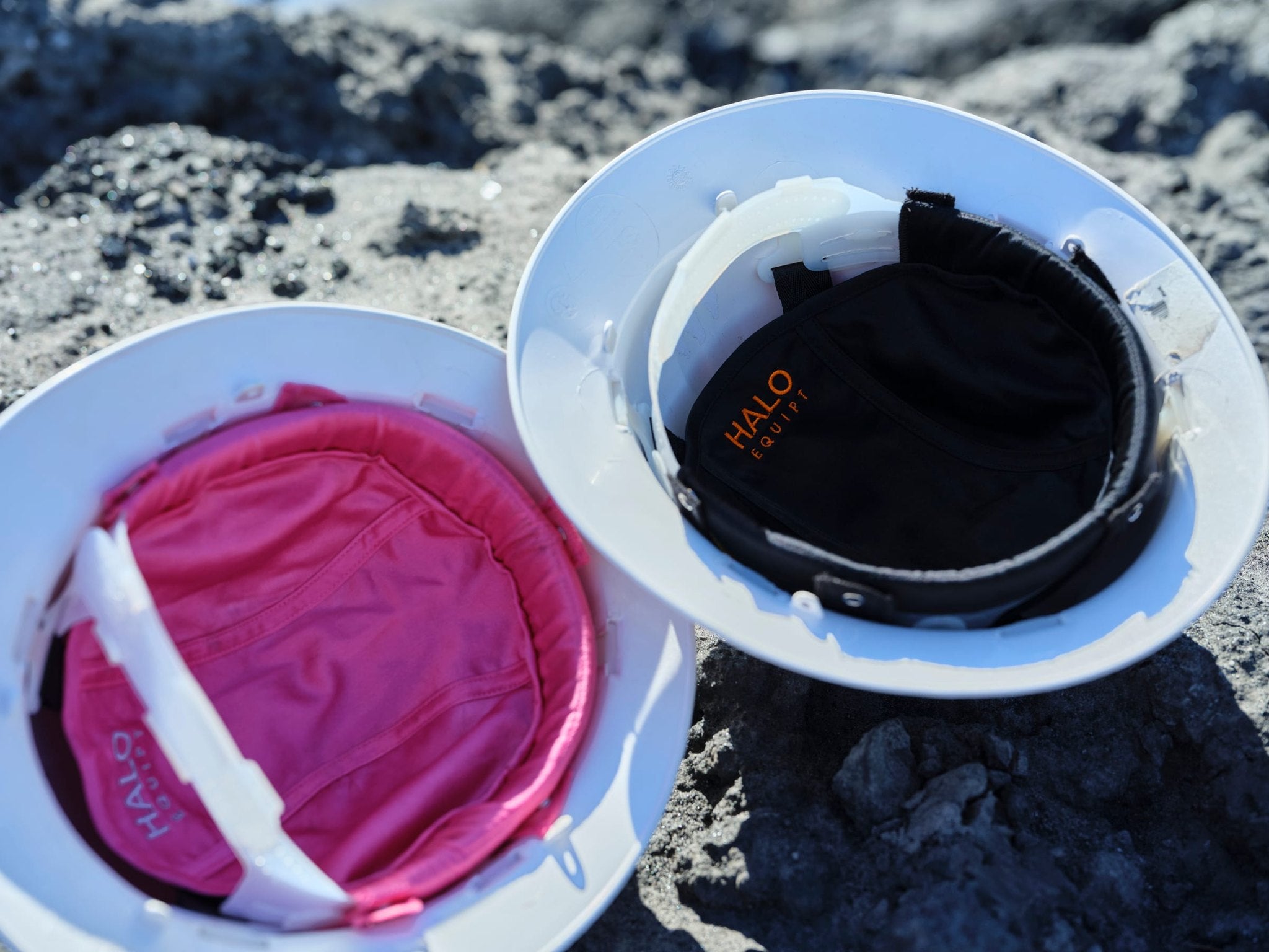 A pink and a black HALO Lid Liner fitted inside two white hard hats, photographed close on rock