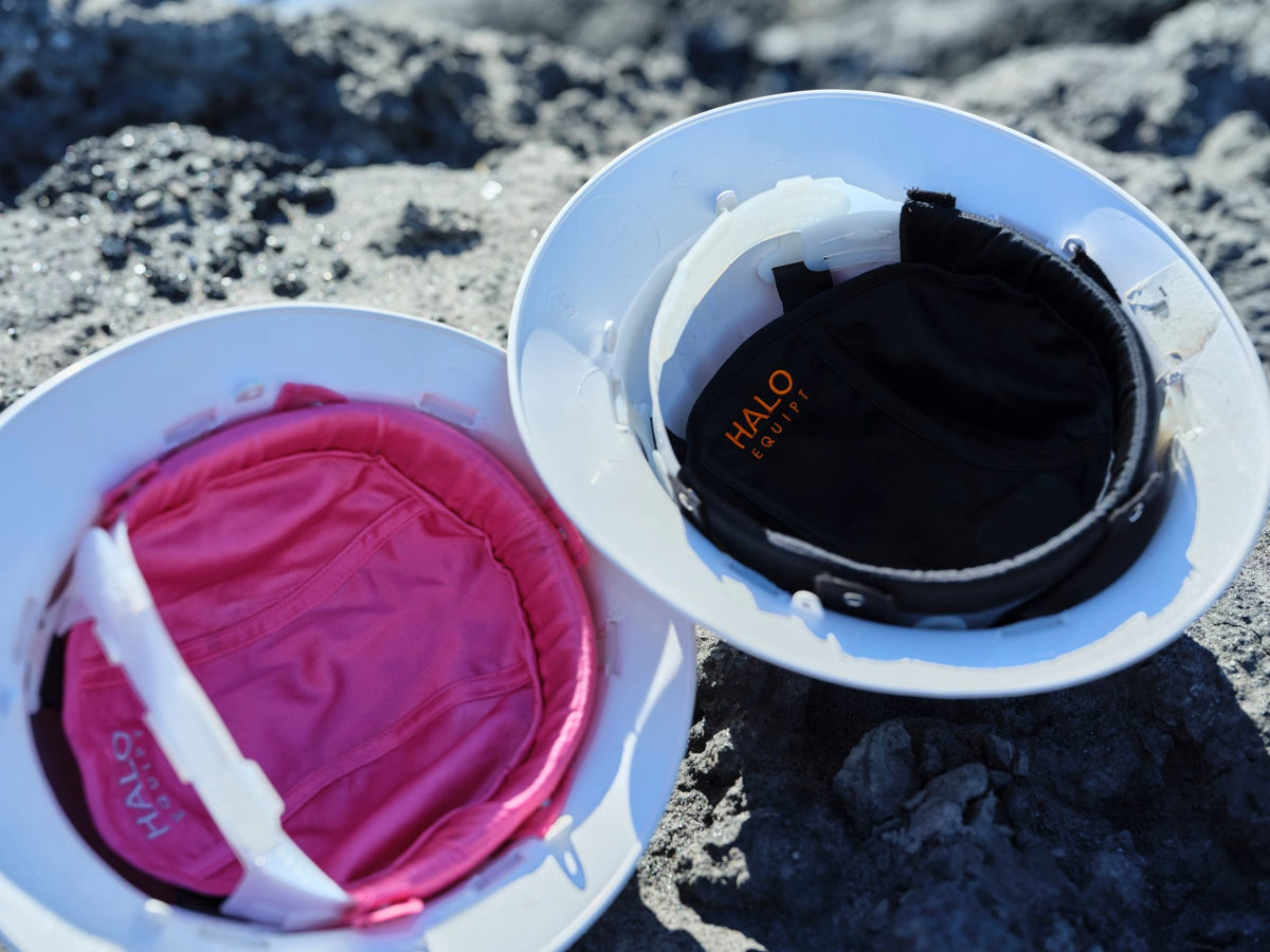A pink and a black HALO Lid Liner fitted inside two white hard hats, photographed close on rock